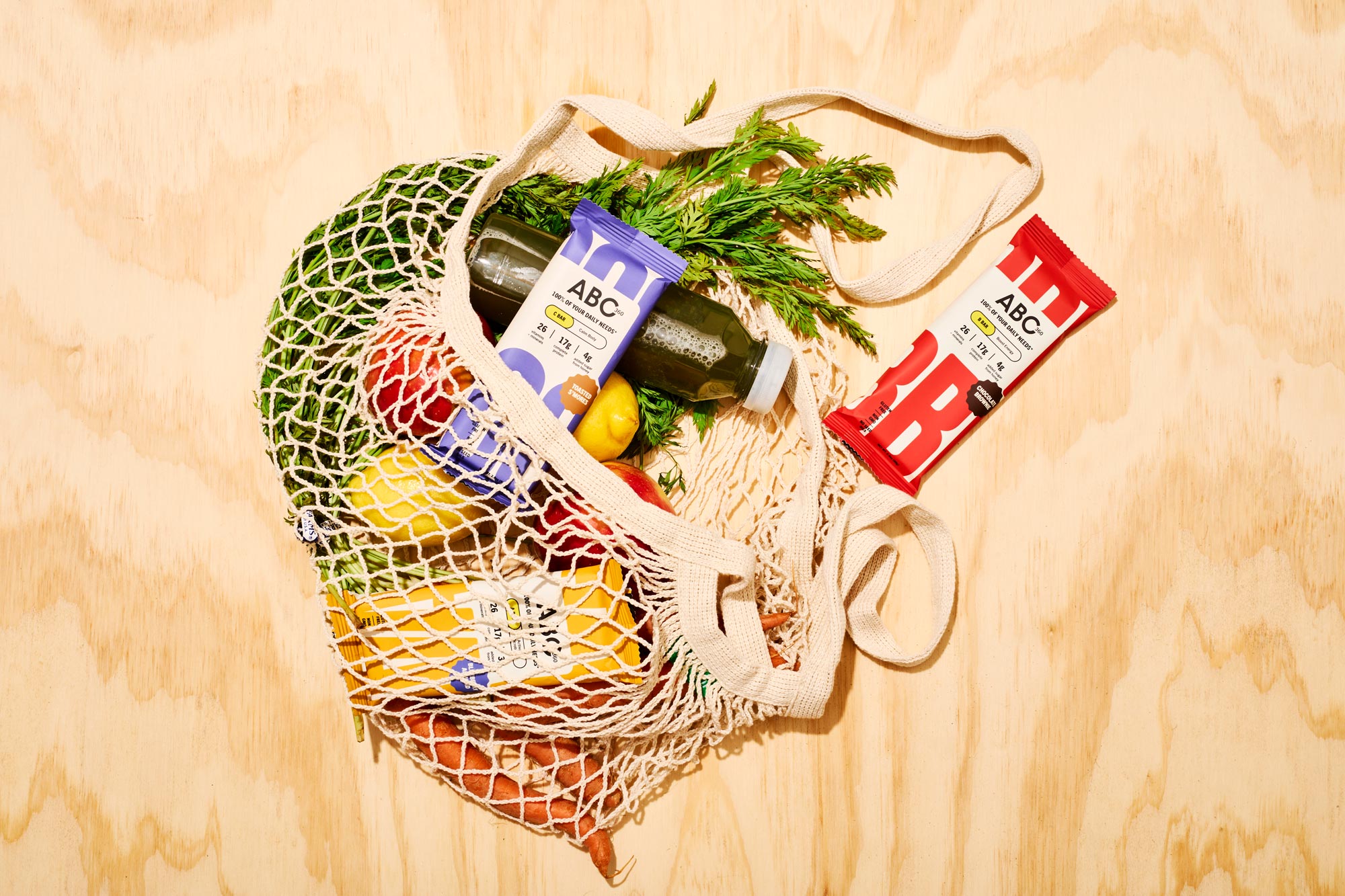 ABC 360 Complete Meal Bars in a reusable mesh grocery bag with fresh produce and a green juice on a wooden surface