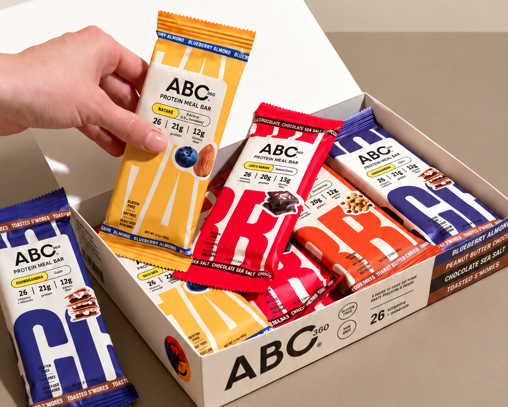 An open ABC 360® Variety Sampler box filled with assorted Protein Meal Bars, with a hand holding up a Blueberry Almond bar.