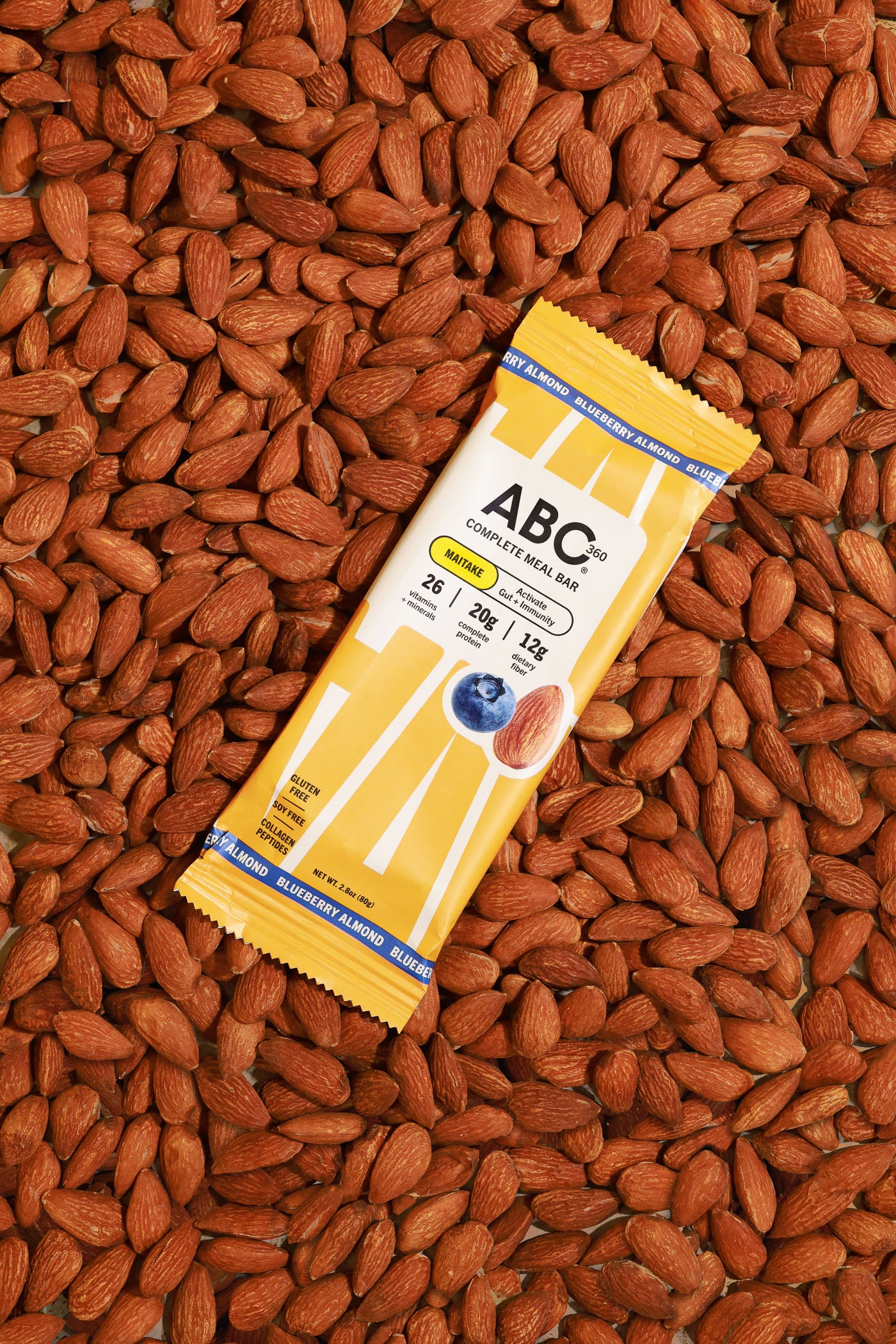 A yellow ABC360BAR.COM Blueberry Almond protein bar wrapper, featuring almond and blueberry images, is set atop a pile of whole almonds, emphasizing its gut health benefits.