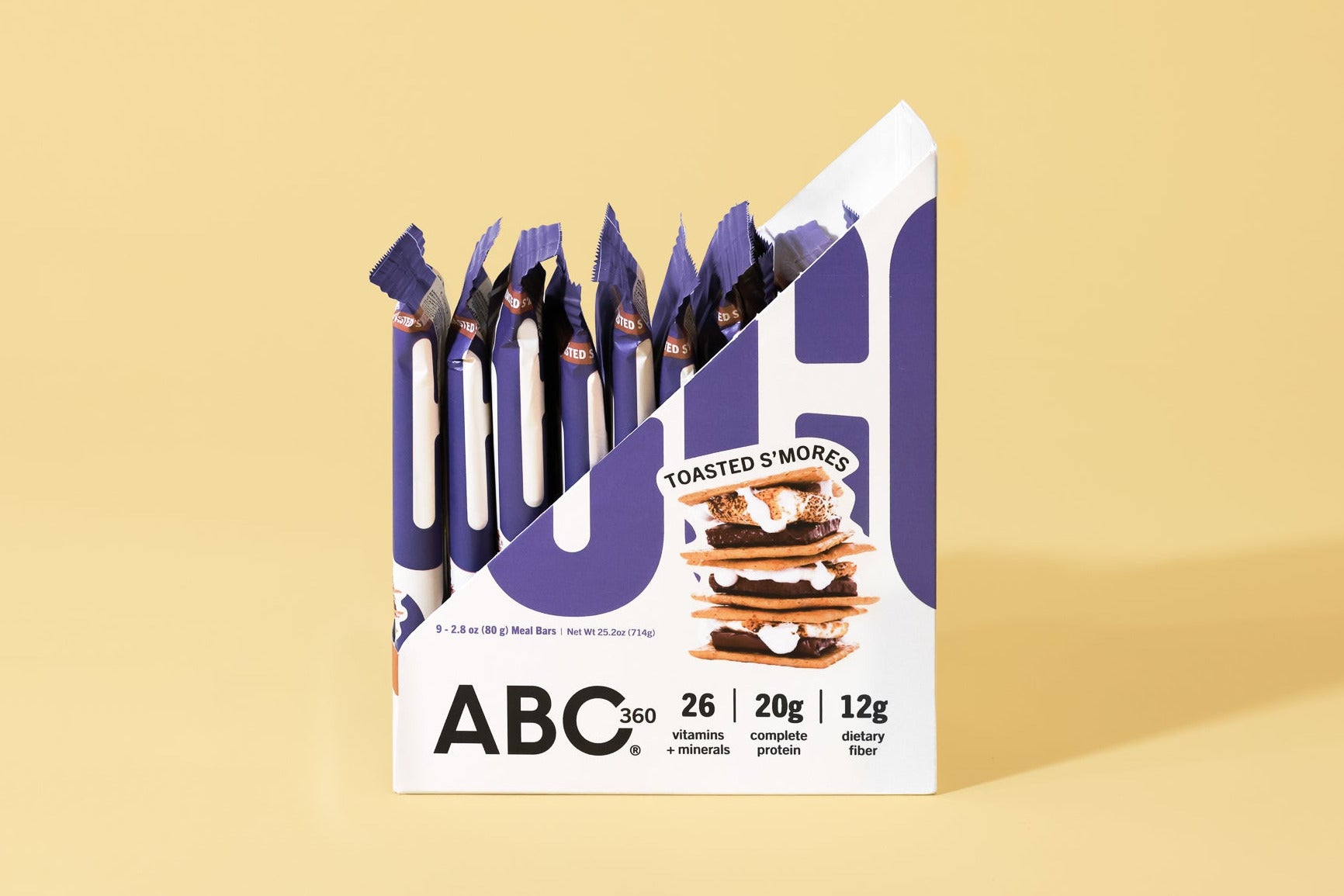An open box of ABC360BAR.COM Toasted S'mores protein bars sits on a yellow background, displaying several bars inside. The packaging highlights 26 vitamins and minerals, 20g protein, and 12g fiber.