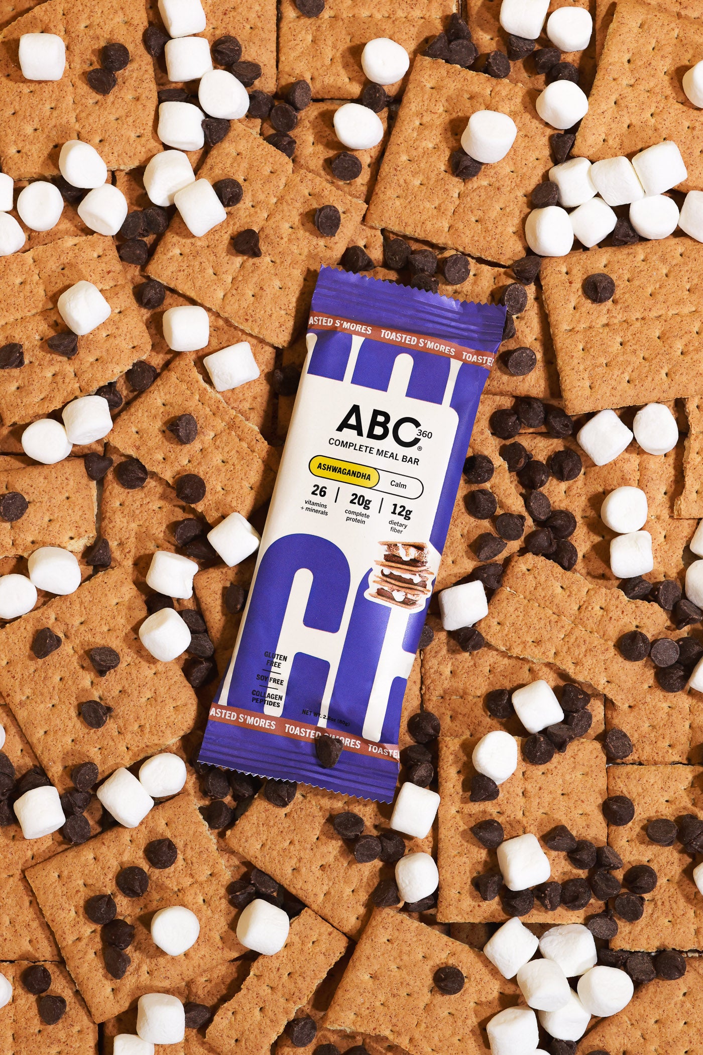 A Toasted S'mores meal bar from ABC360BAR.COM sits on a surface with graham crackers, mini marshmallows, and chocolate chips. The wrapper displays the calming protein bar’s nutrition facts.