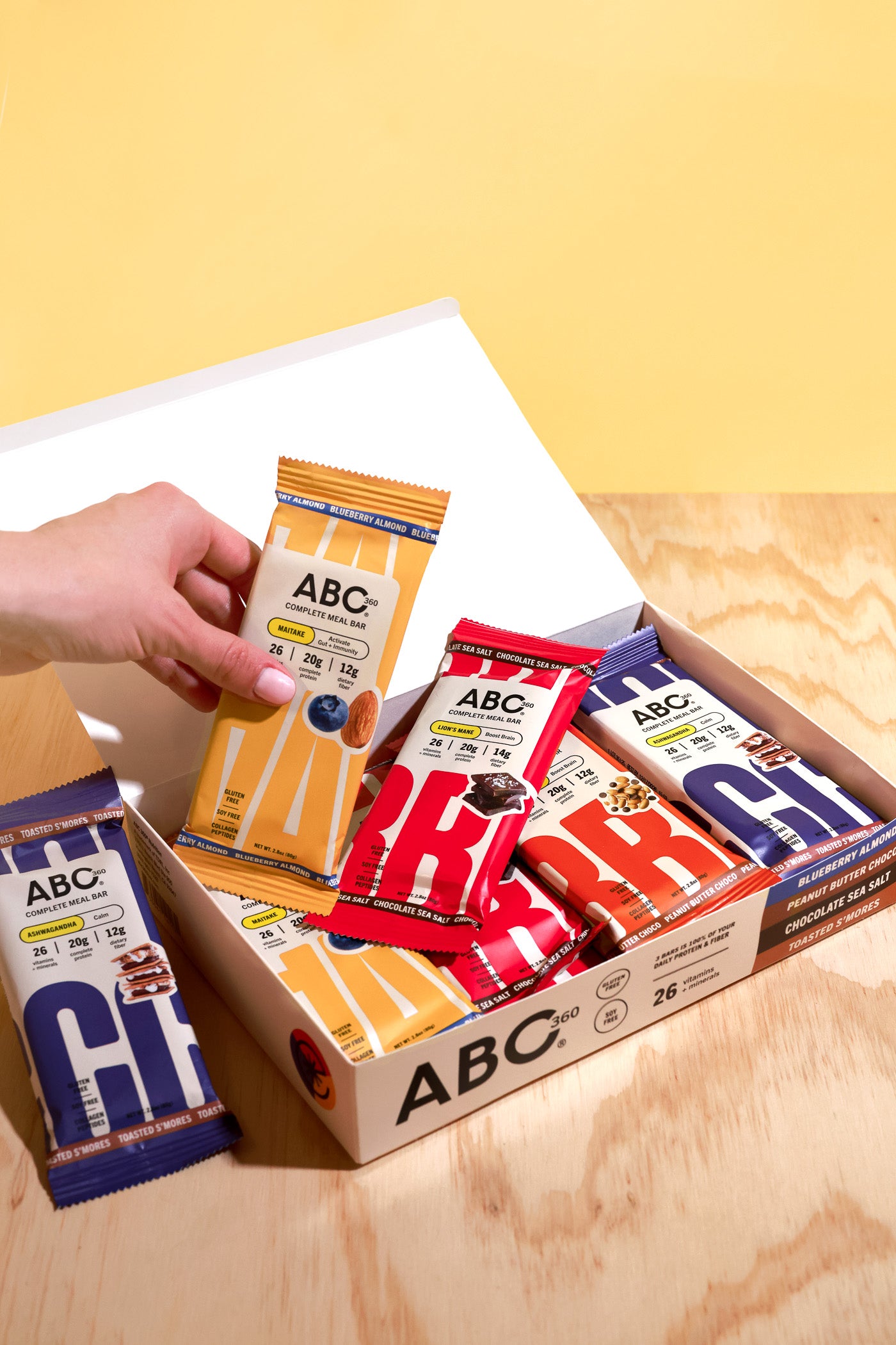 A hand reaches into an open box of assorted ABC360 Bar Giftcards from ABC360BAR.COM in colorful wrappers, placed on a wooden surface with a yellow background.