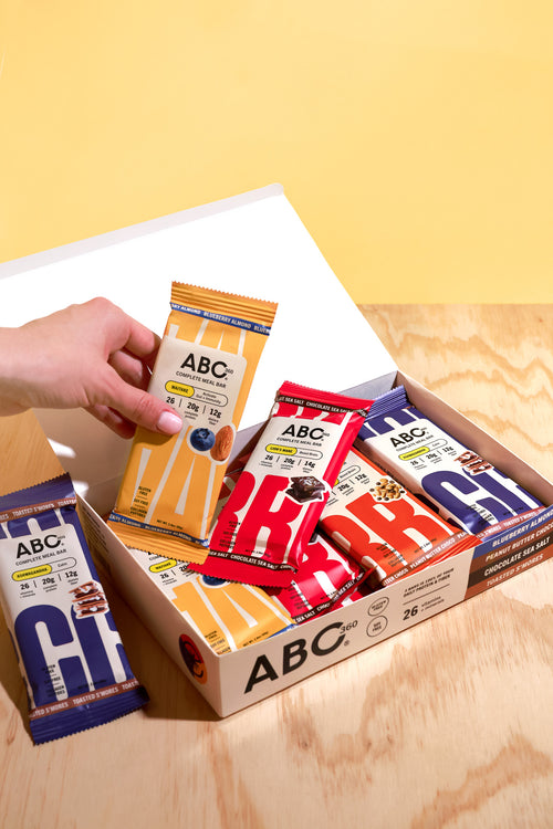 A hand reaches into an open box of assorted ABC360 Bar Giftcards from ABC360BAR.COM in colorful wrappers, placed on a wooden surface with a yellow background.